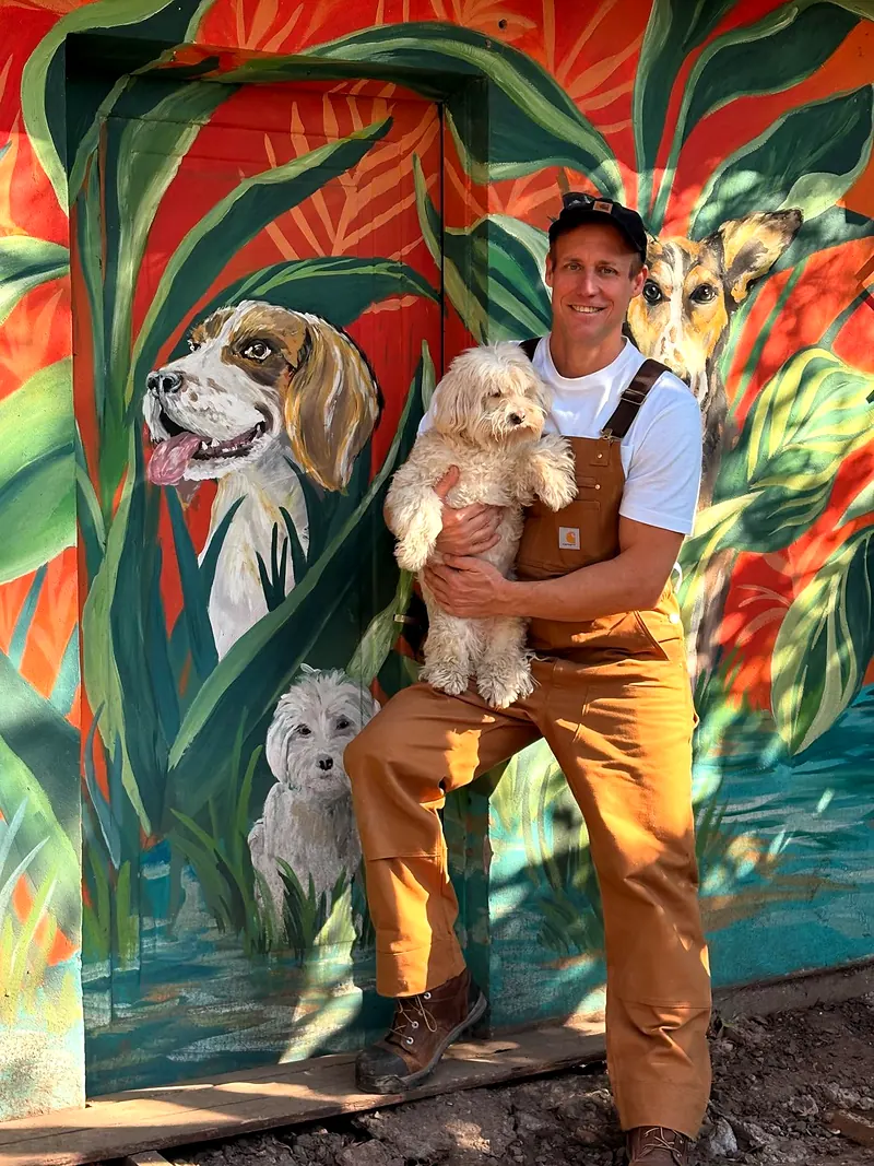 Marcelo con un perro blanco en brazos frente a un mural de perros en la selva